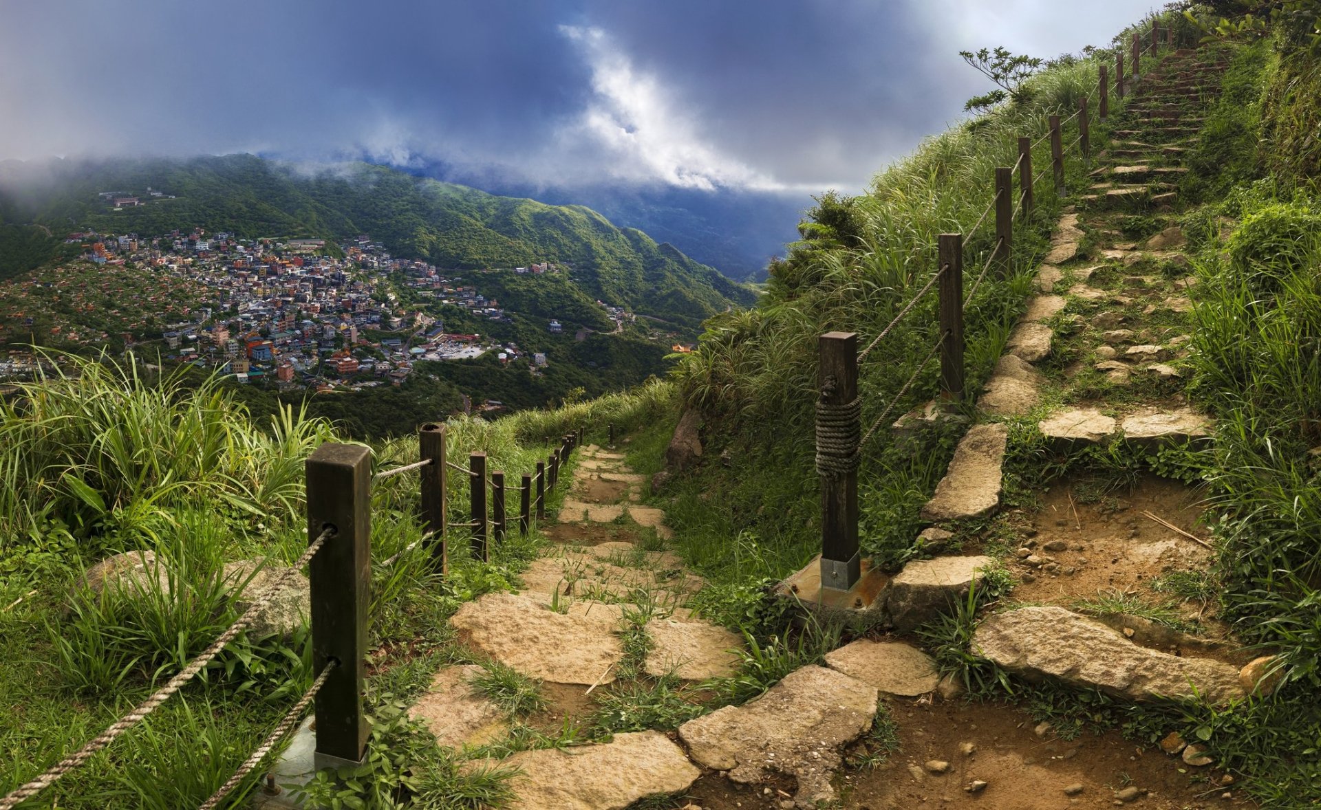 A man-made stone stair path ascends a lush green hillside under a dramatic sky, overlooking a distant village, captured in HD for a PC desktop wallpaper background.