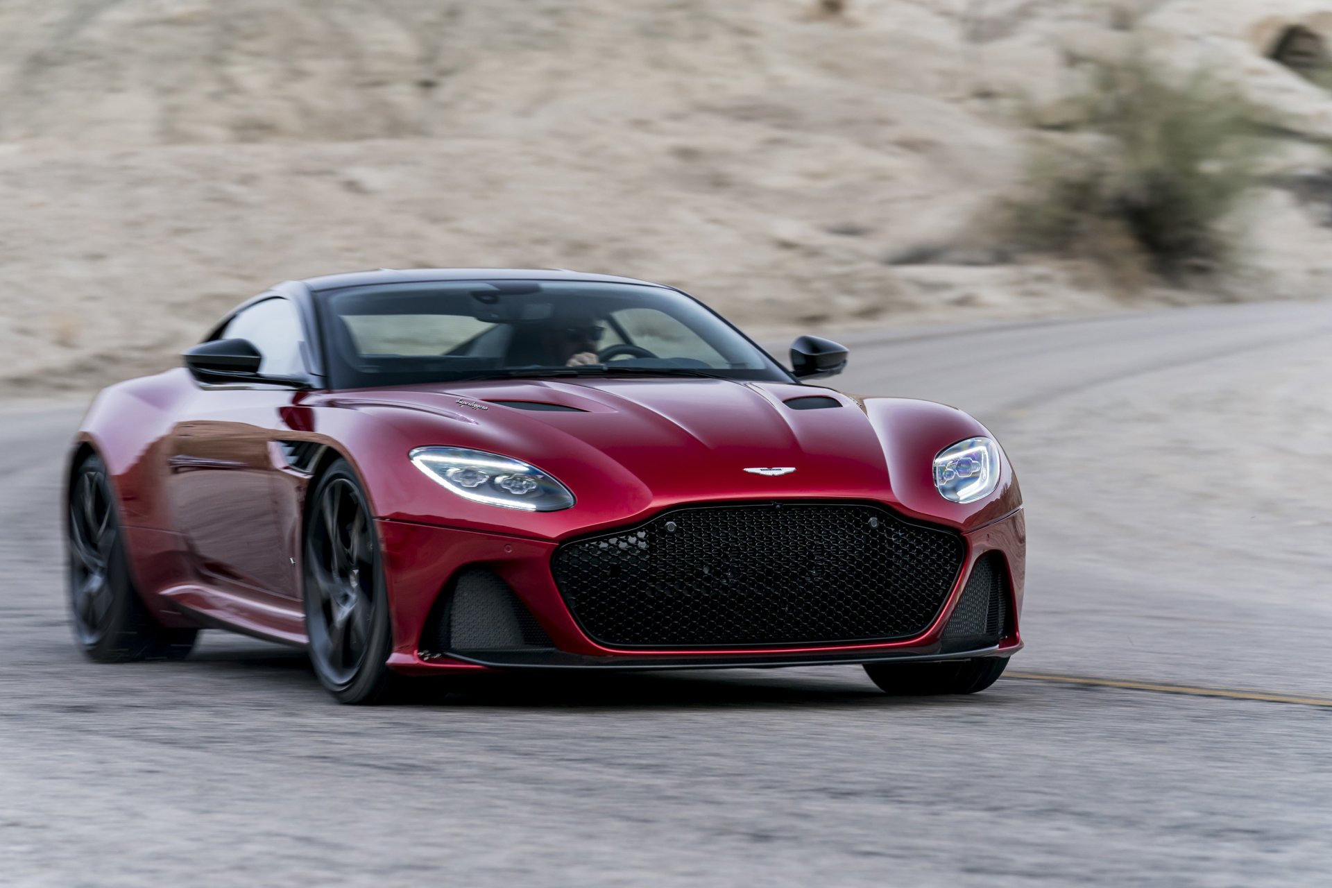 A red Aston Martin DBS Superleggera supercar driving on a winding road, captured in sharp detail for an HD PC desktop wallpaper.