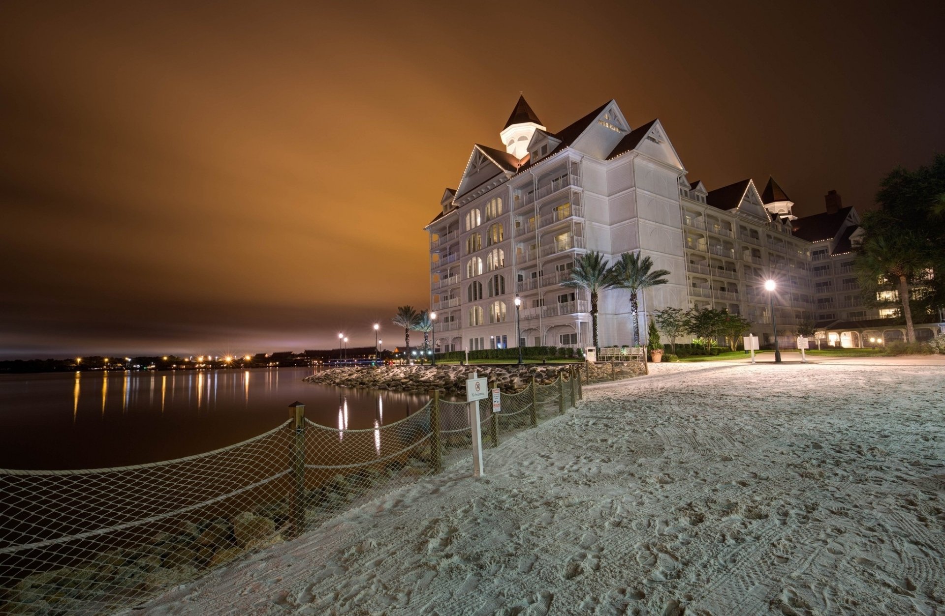 HD desktop wallpaper featuring a man-made hotel illuminated at night by a sandy waterfront with calm waters and a glowing sky.