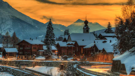 HD desktop wallpaper of a charming Swiss village at sunset, surrounded by snow-covered mountains and rustic buildings. The glowing sky casts a warm light over the serene man-made landscape.
