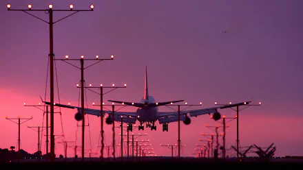 HD wallpaper of a plane landing at twilight, silhouetted against a vibrant pink sky, with runway lights in the foreground.