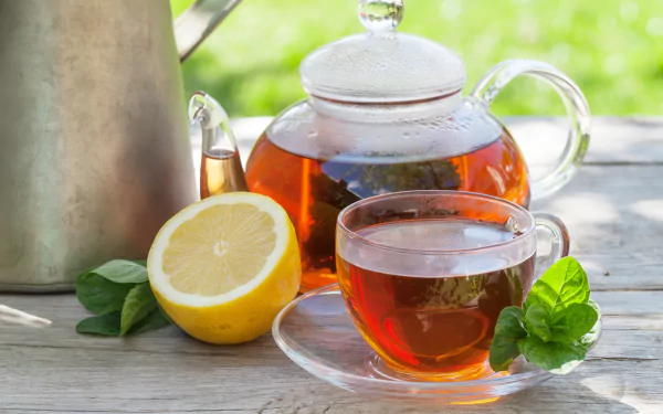 Still-life 2K Quad HD PC desktop wallpaper: glass teapot and teacup of amber tea drink with a halved lemon and mint on a sunlit wooden table.