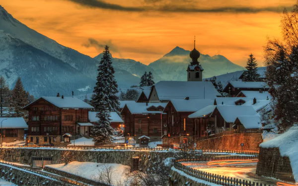 HD desktop wallpaper of a charming Swiss village at sunset, surrounded by snow-covered mountains and rustic buildings. The glowing sky casts a warm light over the serene man-made landscape.