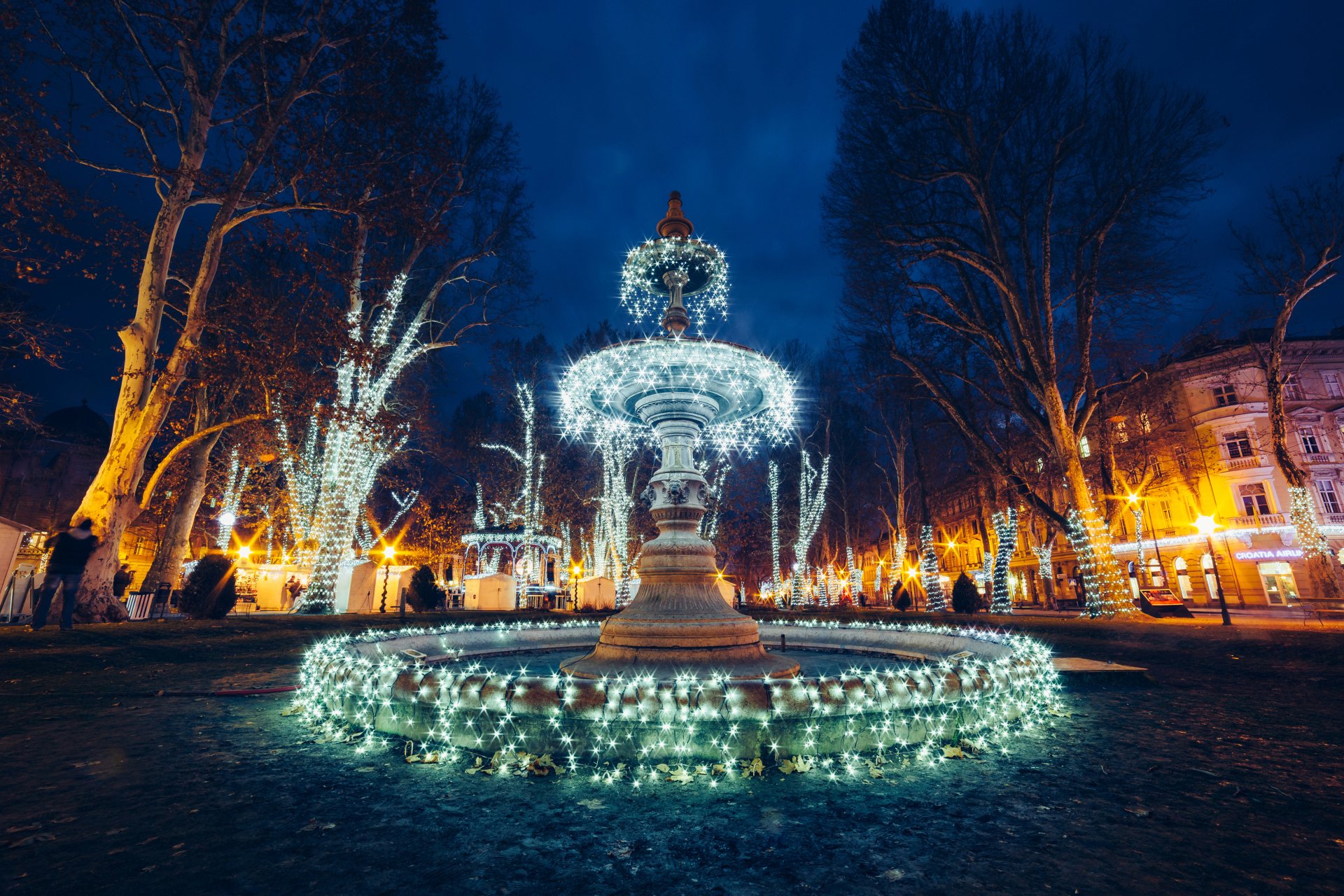 4K Ultra HD PC desktop wallpaper: illuminated man-made fountain at night in a tree-lined square with festive lights.