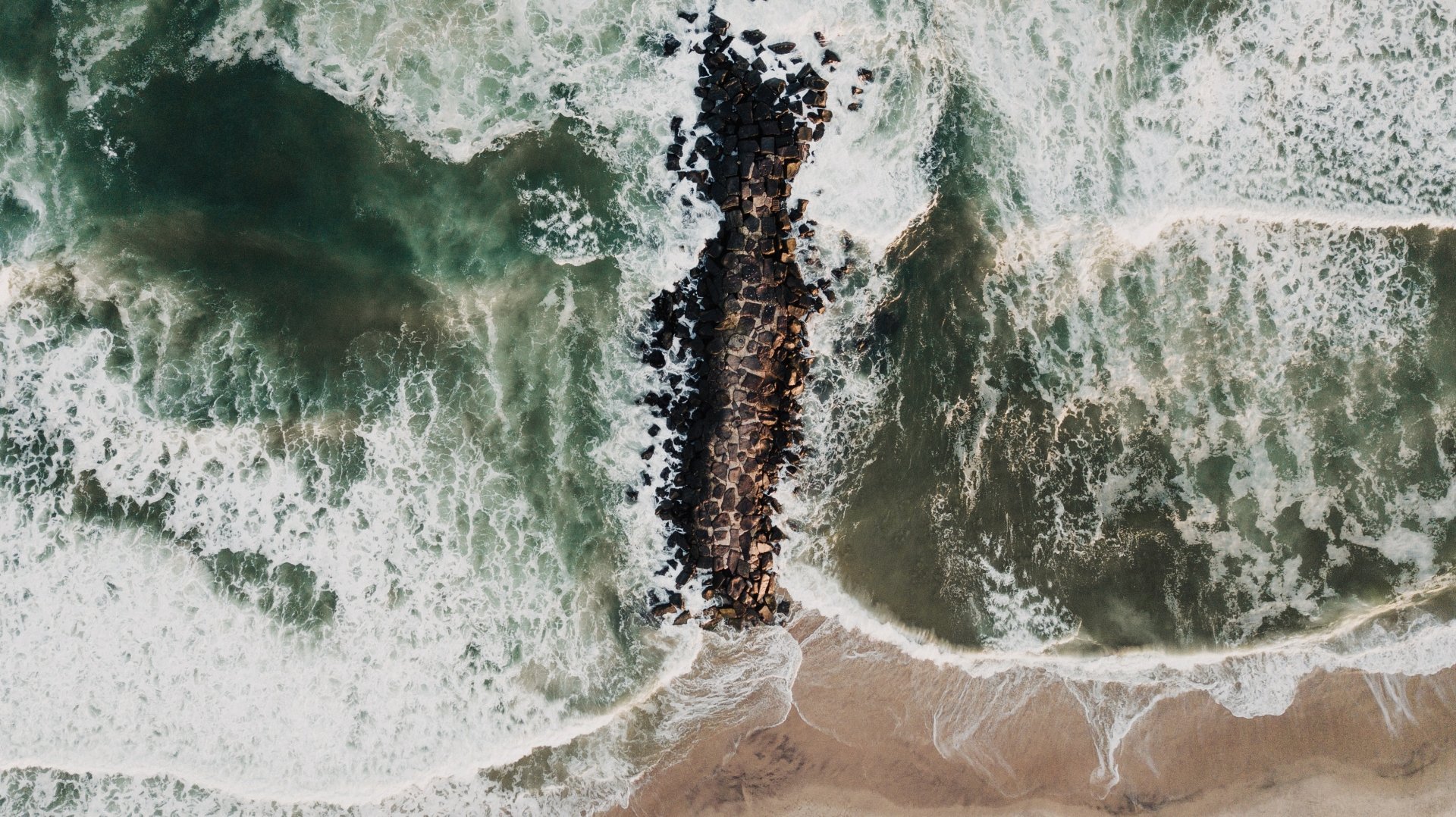 Aerial view of a rocky jetty splitting foamy surf and sandy beach, nature seashore scene — 2K Quad HD PC desktop wallpaper/background.