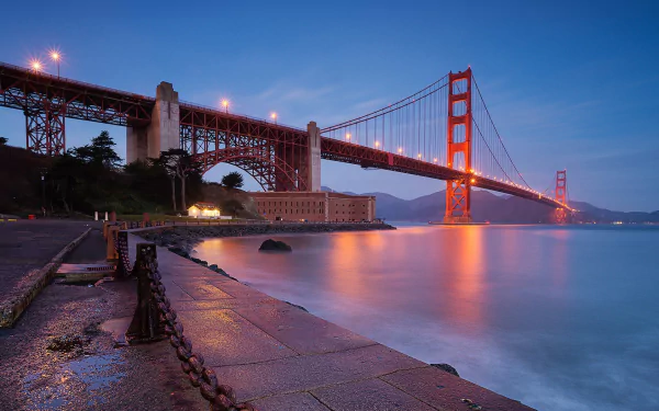 A stunning HD desktop wallpaper showcasing the Golden Gate Bridge at dusk, illuminated and reflecting on calm waters beneath a clear blue sky.