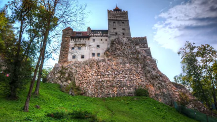 4K Ultra HD image of Bran Castle, a man-made fortress perched on a rocky hill in Transylvania, surrounded by trees under a partly cloudy sky.