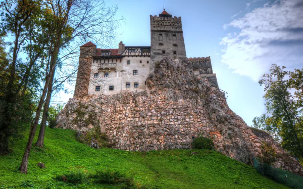4K Ultra HD image of Bran Castle, a man-made fortress perched on a rocky hill in Transylvania, surrounded by trees under a partly cloudy sky.