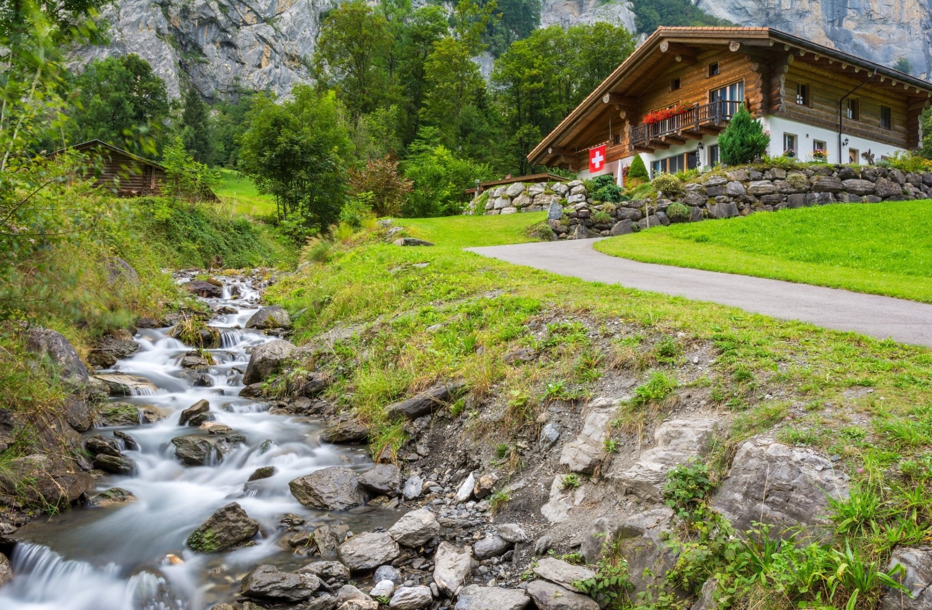 HD desktop wallpaper showcasing a man-made cabin nestled in lush greenery beside a flowing stream, set against a backdrop of rocky cliffs and vibrant natural scenery.