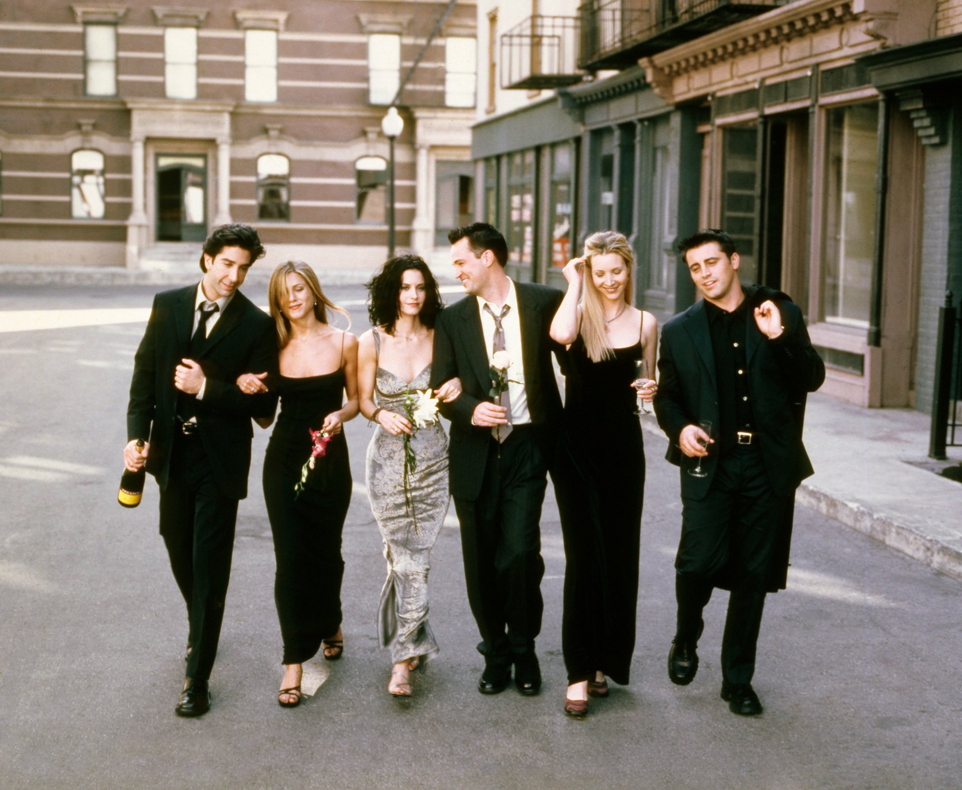 Six friends in evening attire walk arm‑in‑arm down a city street, holding roses and champagne — a stylized HD desktop wallpaper evoking a popular 1990s sitcom ensemble.