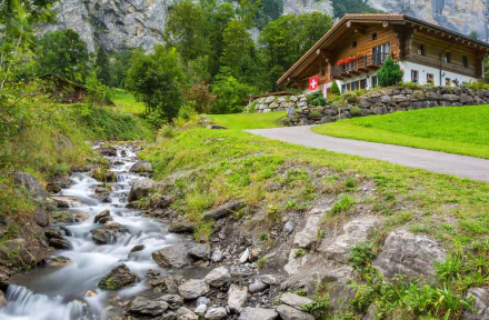 HD desktop wallpaper showcasing a man-made cabin nestled in lush greenery beside a flowing stream, set against a backdrop of rocky cliffs and vibrant natural scenery.