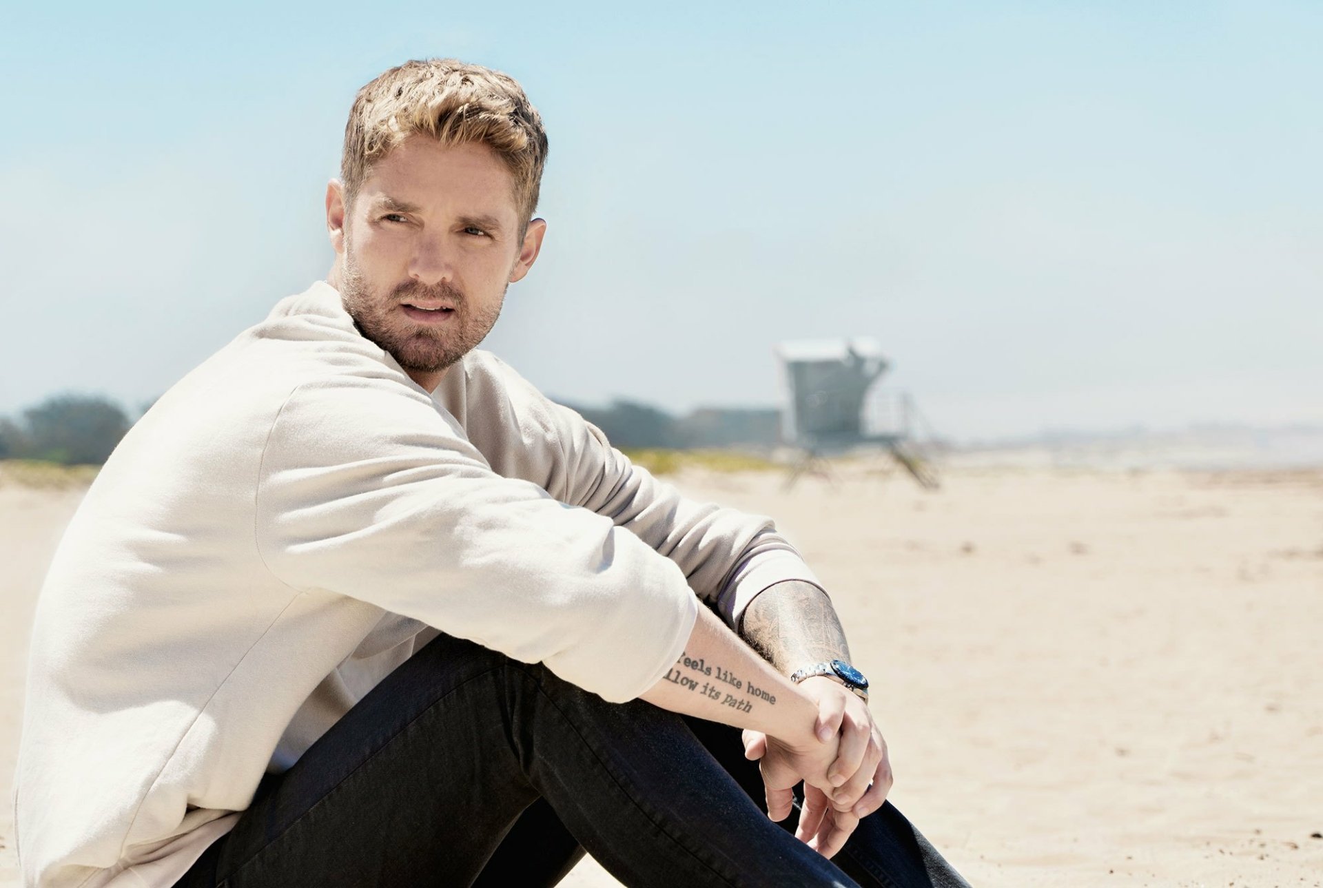 HD desktop wallpaper featuring Brett Young seated on a beach, giving a relaxed, introspective pose with soft focus background.